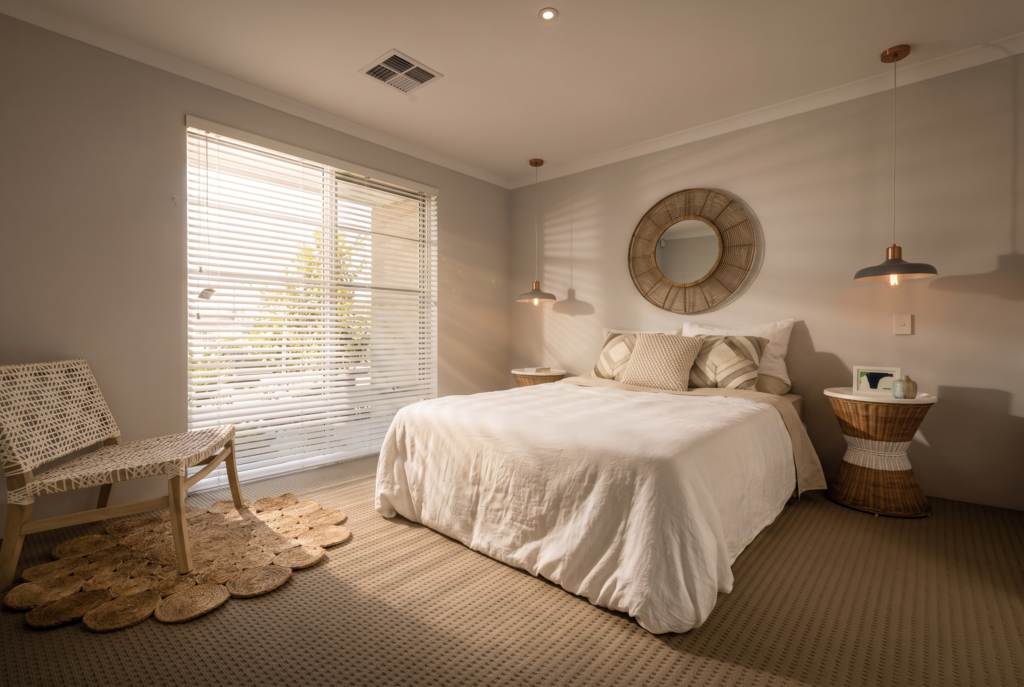 A sunlit bedroom features a bed with white linens, a woven chair, and a round mirror above the headboard.