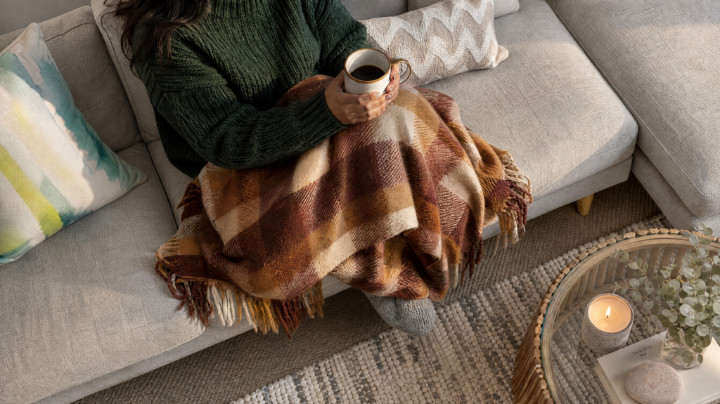 Person in a green sweater holding a mug while wrapped in a plaid blanket on a sofa.