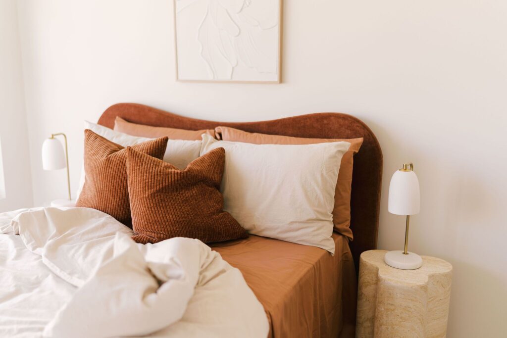 A bed with rust-colored pillows and white bedding is set against a textured headboard.