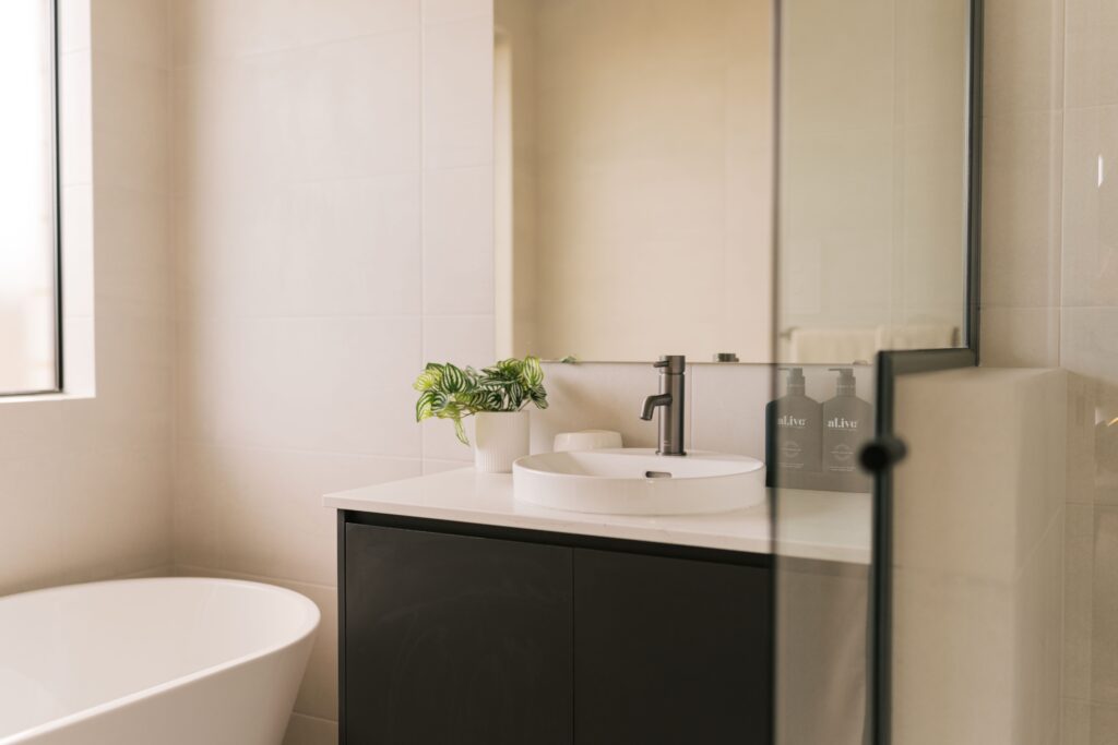 Modern bathroom vanity with a white sink, dark cabinet, and a plant, reflecting two bottles of Alive hand soap.