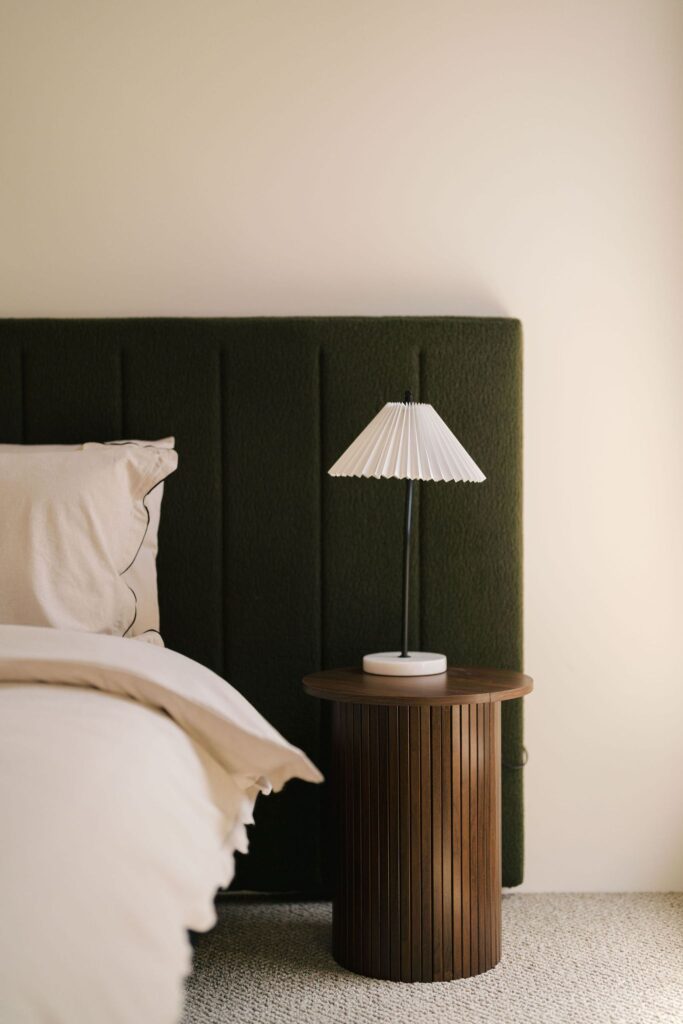 A bedside table with a lamp sits next to a bed with a green headboard.