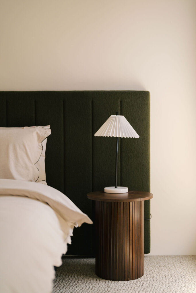A bedside table with a lamp sits next to a bed with a green headboard.