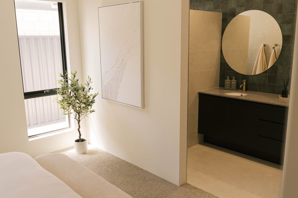 Modern bathroom with dark vanity, round mirror, and green tiled wall, adjacent to a bedroom with a potted plant.