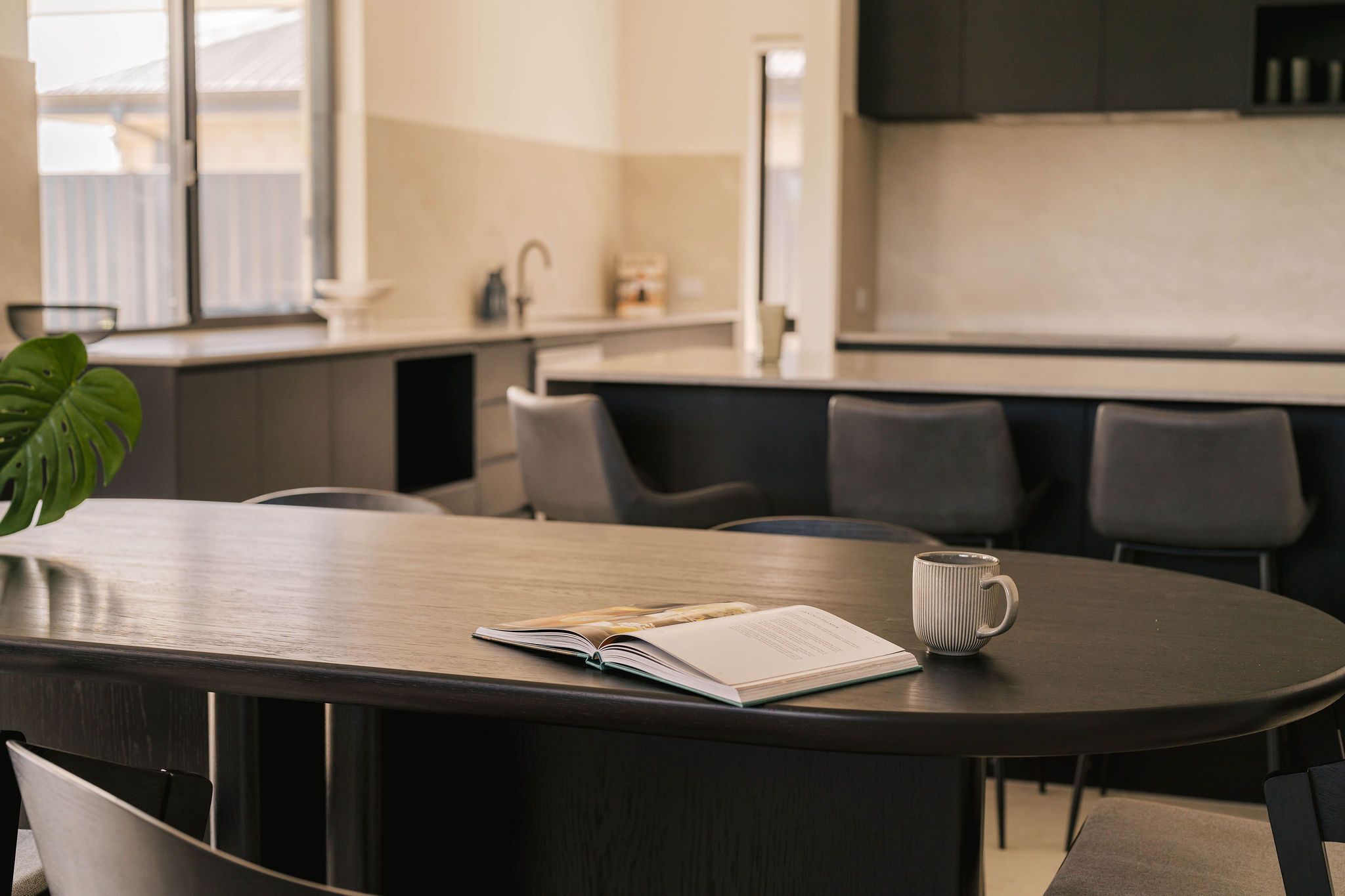 Open book and mug on a dark oval dining table in a modern kitchen.