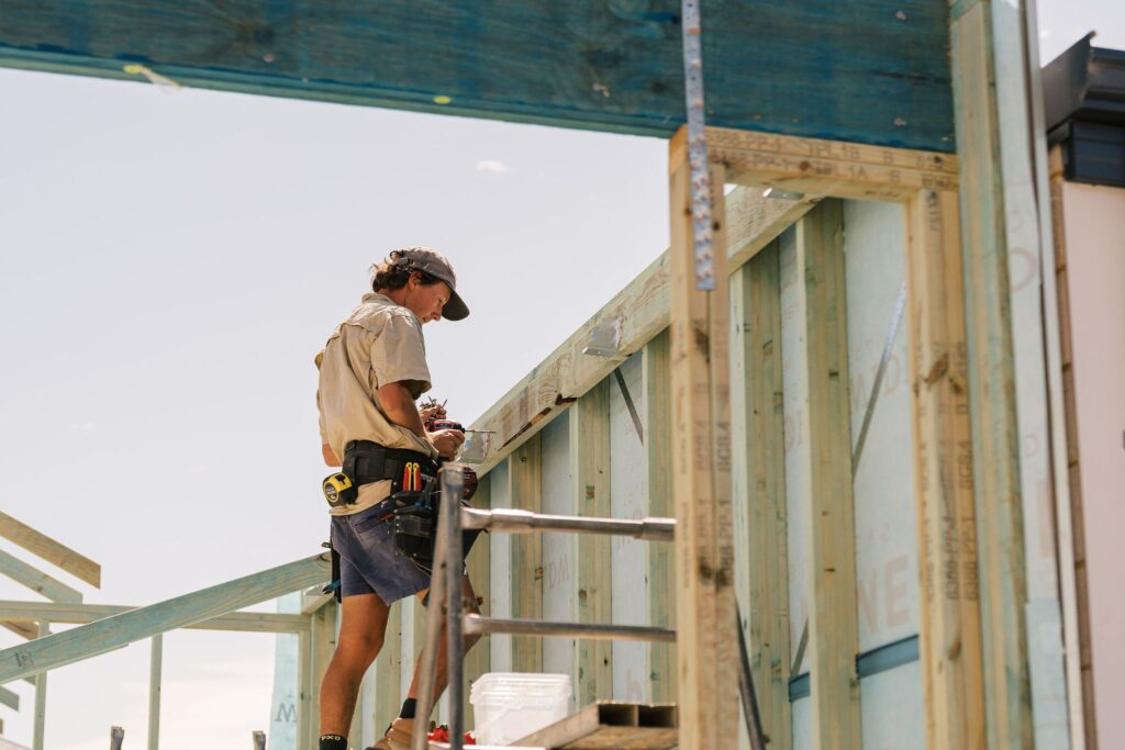 A builder uses a drill to attach a metal bracket to the wooden frame of a new house.