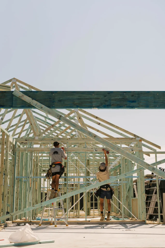 Two builders work on the timber frame of a new house under a clear sky.