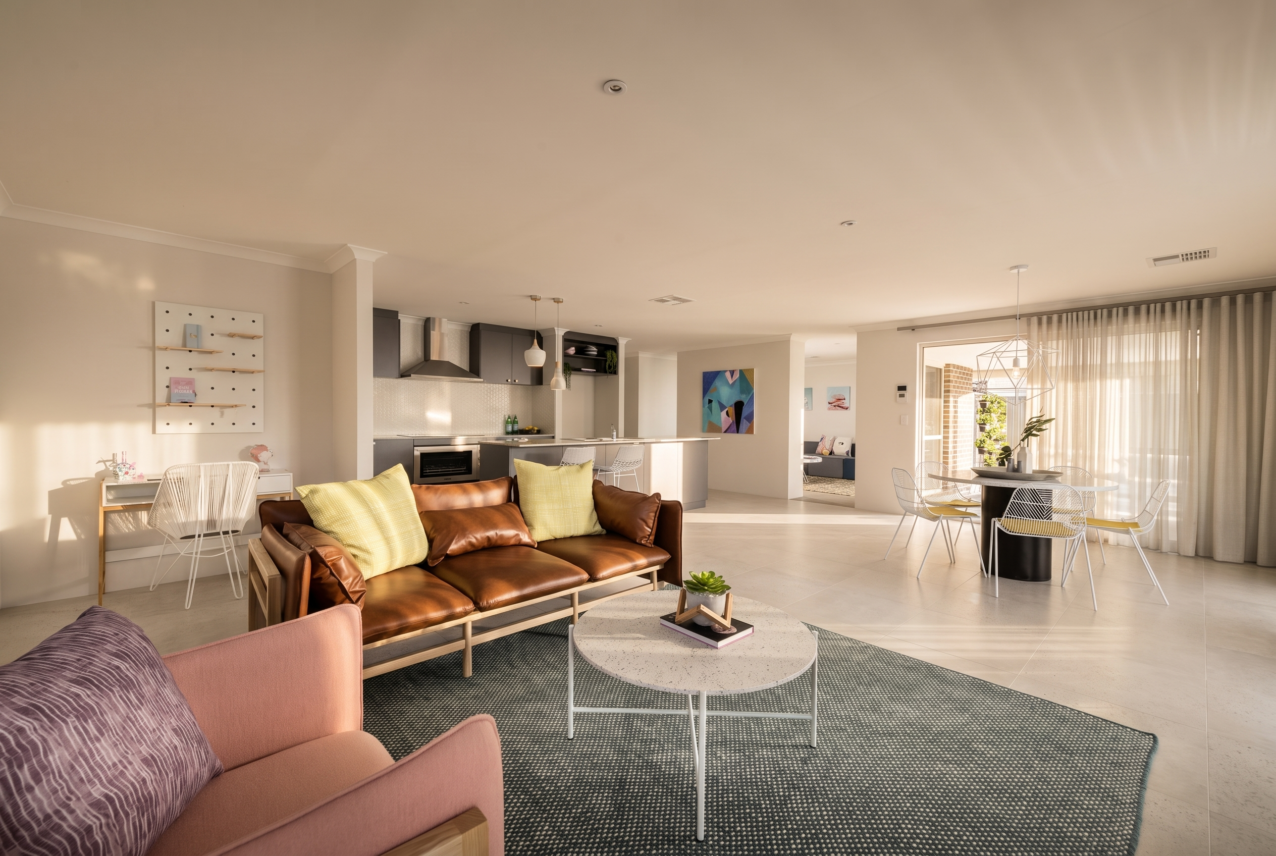 A spacious, sunlit open-plan living area with a brown leather sofa, pink armchair, and modern kitchen.