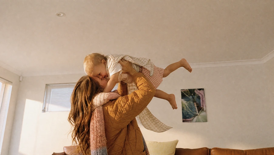 A mother lifts her baby high in the air in a bright, modern living room.