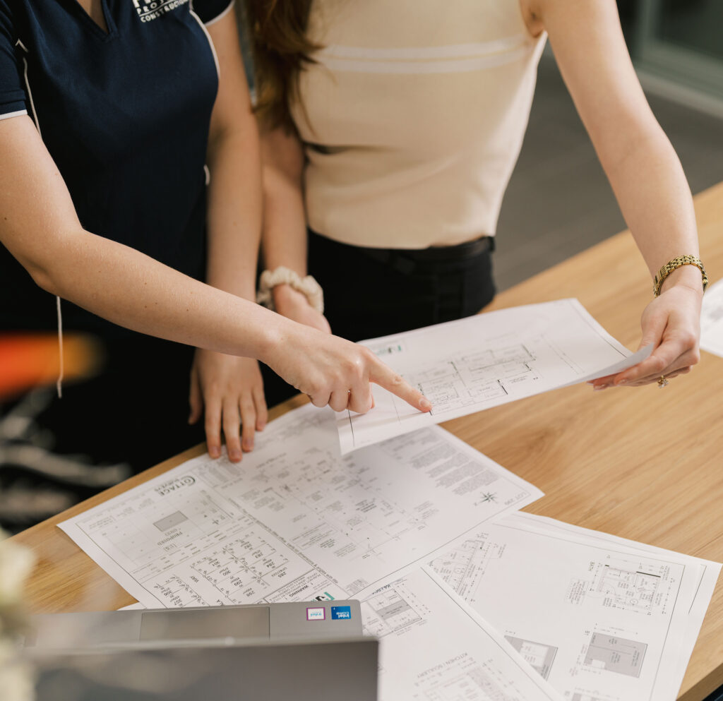 Two women review architectural plans on a wooden desk, one pointing to a detail.