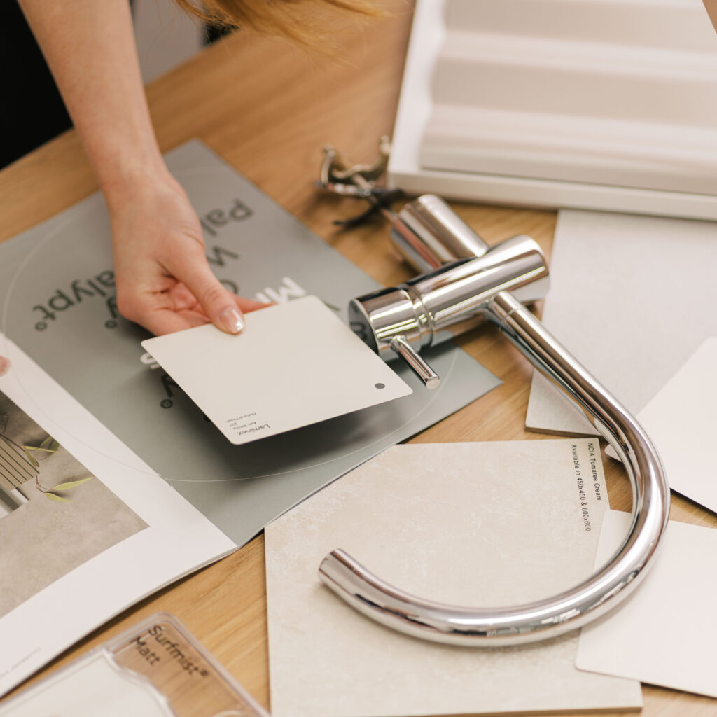 A hand holds a light beige paint swatch over a selection of tiles and a chrome faucet.