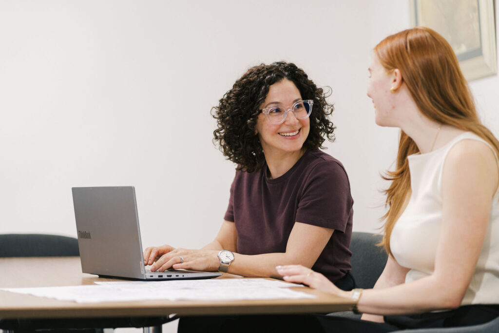 Two women smiling and looking at a laptop on a table.