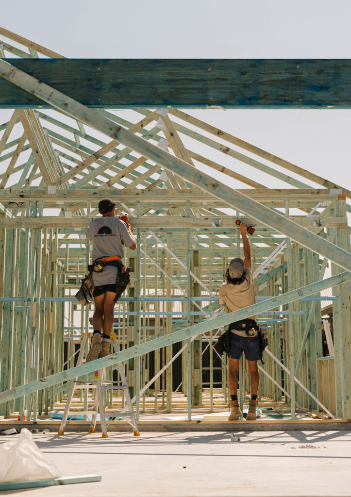 Two builders work on the timber frame of a new house under construction.